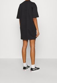 Svart oversized t-shirt klänning, korta ärmar, rund halsringning, bomullstyg. Bärs med svarta sneakers och vita strumpor, står på en neutral yta.