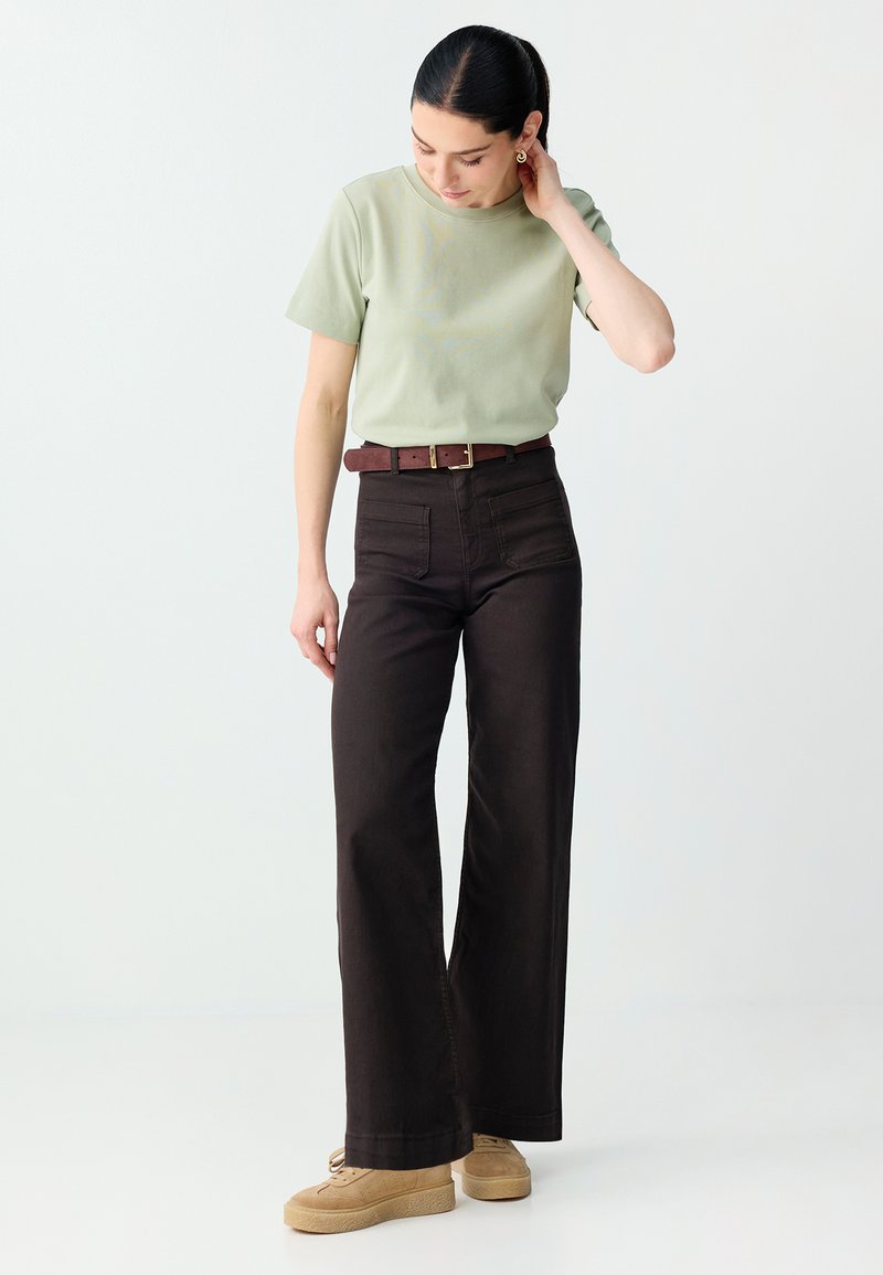 Femme en chemise à manches courtes vert clair, pantalon large noir taille haute, ceinture marron, chaussures à plateforme beiges, regardant vers le bas avec la main près de l'oreille.