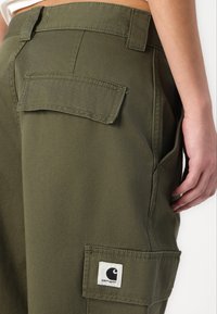 Olivgrüne Cargo-Hosen mit lockerer Passform, ausgestattet mit zwei Gesäßtaschen und einem Markenpatch an der Seite. Robuste, glatte Stofftextur.