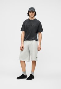 Νέος άνδρας που φοράει ξεθωριασμένο μαύρο μπλουζάκι, ανοιχτόχρωμο γκρι μακρύ σορτς, γκρι κάλτσες, μαύρα αθλητικά παπούτσια και μαύρο καπέλο του μπέιζμπολ, στέκεται σε λευκό φόντο.