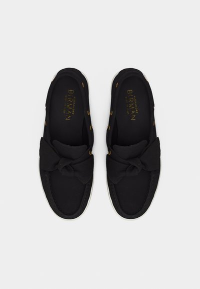 Γυναικεία παπούτσια τύπου slip-on σε μαύρο χρώμα με μεγάλα υφασμάτινα φιόγκους στην κορυφή, στρογγυλές μύτες και λευκές σόλες, εμφανιζόμενα από ψηλά σε λευκό φόντο.