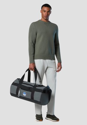 Uomo con maglione verde e pantaloni bianchi che tiene una borsa da viaggio cilindrica grigia e nera con logo su sfondo chiaro.