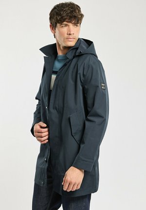 Homme portant un imperméable à capuche bleu foncé par-dessus un pull rayé, posant avec les mains près de l'avant du manteau devant un fond uni.