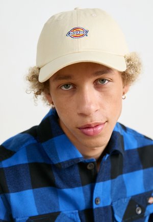 Cappellino da baseball beige con logo ricamato, indossato da una persona in una camicia di flanella a quadri blu e nera. Tessuto morbido, visiera curva.