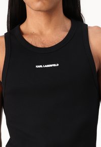 Črn rebrast top brez rokavov z okroglim ovratnikom in napisom "KARL LAGERFELD" v beli barvi na sredini. Material se zdi tesno priležen.