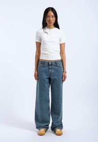 Hvid kortærmet tætsiddende top, lyseblå oversized denimjeans, gule snørebåndssko og enkel smykker mod en ensfarvet baggrund.