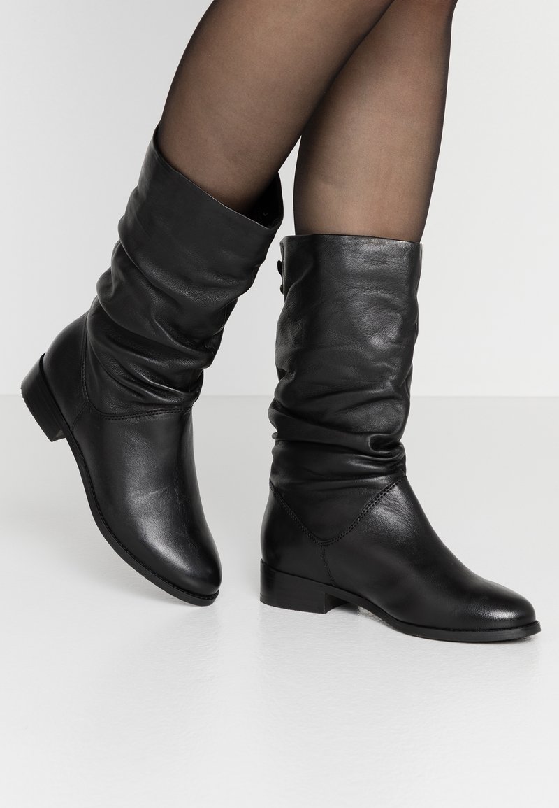Gambe in collant neri trasparenti che indossano stivali neri in pelle morbidi a metà polpaccio con tacco basso su un pavimento bianco.