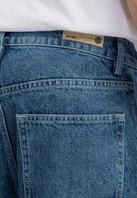Jeans in denim blu con tasca posteriore e un'etichetta con la scritta "EST. 1992". Il design è completato da cuciture visibili e un tessuto texturizzato.