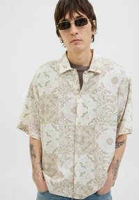 Camisa de manga corta con botones en beis claro, con un patrón floral texturizado, que presenta un cuello y un corte relajado.