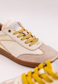 Sneaker beige e color sabbia realizzati in materiali scamosciati e in pelle testurizzati, con lacci gialli testurizzati e una forma leggermente arrotondata.