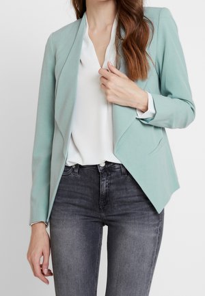 Woman wearing a pastel green blazer over a white blouse and faded black jeans, holding blazer lapel with one hand.