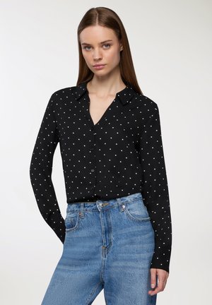 Donna che indossa una maglietta nera a maniche lunghe con pois bianchi, infilata in jeans blu a vita alta, in piedi davanti a uno sfondo semplice.