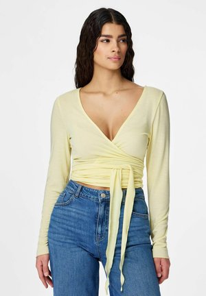 Žena s dlhými tmavými vlasmi má na sebe svetložltý zavinovací top s viazaním vpredu a vysoko pásované modré džínsy, pozerá sa mierne do strany.