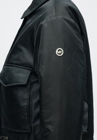 Μαύρο μπουφάν από λείο, γυαλιστερό υλικό με γιακά, τσέπη ραμμένη επάνω και ασημένιο κυκλικό λογότυπο στο αριστερό μανίκι.