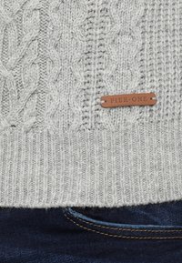 Maglione grigio chiaro lavorato a maglia con motivi a treccia e un'etichetta in pelle marrone con la scritta "PIER ONE" sopra un paio di jeans in denim blu scuro.