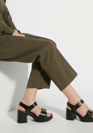 Zwarte leren platform sandalen met een gespaccent, open teen en dikke blokhak, gecombineerd met lange olijfgroene broek.
