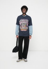 Tricou grafic bleumarin cu inscripția "CROWN JEWEL KINGS LOS ANGELES", stratificat cu mâneci lungi de culoare bleu deschis. Pantaloni negri și adidași eleganți.