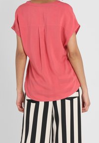 Blusa de coral con mangas cortas, material suave, detalle de pliegue en la parte posterior y dobladillo curvado; combinada con pantalones de pierna ancha a rayas blancas y negras.