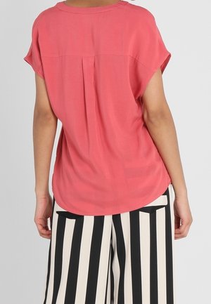 Blusa di corallo con maniche corte, materiale morbido, dettaglio a piega sul retro e orlo curvo; indossata con pantaloni a gamba larga a righe bianche e nere.