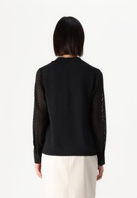 Musta toppi, jossa on kiinteä kangas ja pitkät, läpinäkyvät hihat, joilla on chevron-kuvio. Pyöreä kaula-aukko ja nappikainalot. Yksinkertainen takatyyli.