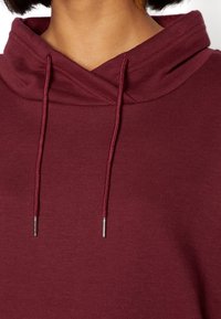 Sudadera con capucha en color burdeos que presenta un escote cruzado, un tejido texturizado y cordones ajustables con puntas de metal plateado.