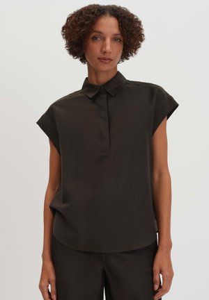 Frau mit kurzen lockigen Haaren, die ein lockeres schwarzes kurzärmeliges Hemd mit Kragen und schwarze Hose trägt, steht vor einem einfarbigen weißen Hintergrund.