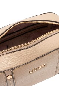 Beżowa teksturowana skórzana torba ze złotym zamkiem błyskawicznym i ozdobami. Wnętrze wyściełane wzorzystą podszewką. Na boku wytłoczone logo "LIUJO".
