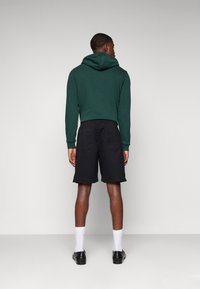 Sudadera verde con una gran capucha, combinada con pantalones cortos negros que tienen bolsillos laterales. El modelo está de espaldas al espectador, mostrando un diseño sencillo.