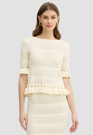 Donna che indossa un completo crema di top e gonna lavorati a maglia con motivi traforati, orlo e maniche arricciate e orecchini a cerchio dorati.