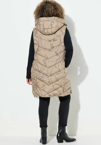 Beige Steppweste mit abstrakter Gesichts-Linienkunst, ausgestattet mit Kapuze, gesteppter Textur und seitlichen Taschen. Kombiniert mit schwarzen Jeans und Stiefeln.