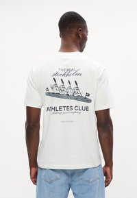Tricou alb din bumbac, cu un imprimeu grafic pe spate cu figuri de vâslire și text, croială lejeră, mâneci scurte și guler rotund.