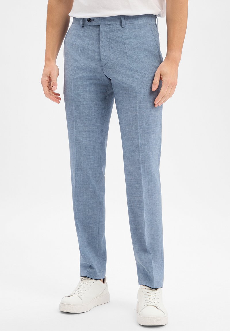 Mann trägt hellblaue, schmal geschnittene Anzughose mit Bügelfalte, kombiniert mit weißen Turnschuhen und einem weißen Hemd, steht vor weißem Hintergrund.