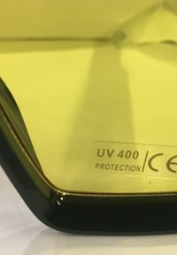 Occhiali da sole neri con etichetta di protezione UV 400, caratterizzati da una finitura lucida e un design angolare, su uno sfondo giallo.