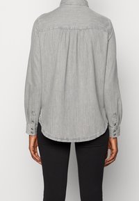 Grå denimskjorte med knaplukning fortil, foldet krave, lange ærmer og søget detaljer bagpå. Rundet kant med sideslids.