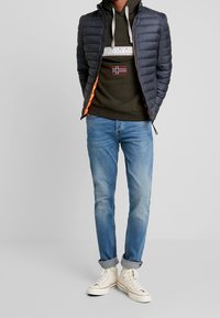 Giacca trapuntata blu navy con fodera arancione, felpa verde oliva con un dettaglio grafico e bandiera, jeans azzurri chiari e sneakers alte bianche.