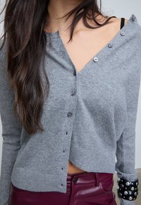 Vrouw in een grijze knopencardigan die van één schouder glijdt, met een zwarte gespijkerde armband en hooggewaisted kastanjebruine leren broek.