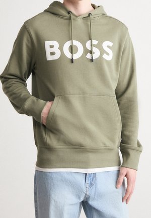Personne portant un sweat à capuche vert olive avec le texte blanc "BOSS", un jean bleu clair et une main dans la poche du sweat à capuche, sur un fond uni.