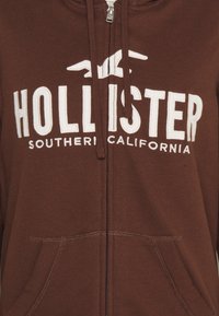 Brun zip-up hoodie med en stor vit "HOLLISTER"-logotyp och texten "SOUTHERN CALIFORNIA", två framfickor och ribbstickade muddar.