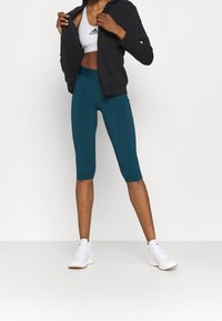 Tyrkysové legíny s vysokým pasem a černá krátká mikina na zip, doplněné bílými sportovními botami, prezentované na jednoduchém pozadí.