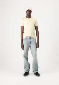 T-shirt jaune clair, jean bleu décontracté et baskets noires. Une casquette blanche est portée. Design simple sans motifs ni logos visibles.