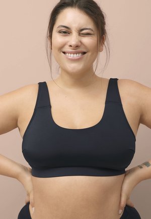 Sort sports-bh med brede stropper og scoop-udskæring, lavet af glat, strækbart stof. Båret af en smilende model foran en neutral baggrund.