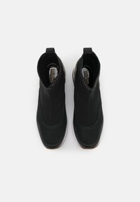 Zapatos negros tipo slip-on con parte superior de malla, punta redondeada y suela de goma. Presentan una lengüeta para tirar y un logotipo en el cuello. Superficie plana y texturizada.