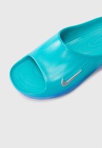 Tongs Nike turquoise avec une ouverture à l'avant, texture lisse et brillante, et un accent logo argenté sur le côté. Elles présentent une semelle intérieure moulée et une semelle bleue.