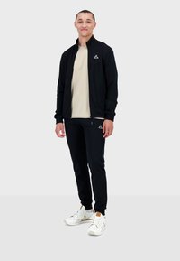 Giacca nera con zip e pantaloni neri abbinati a una t-shirt beige chiaro. Sneakers bianche con dettagli e loghi dorati. Caratterizzata da un design slim fit.