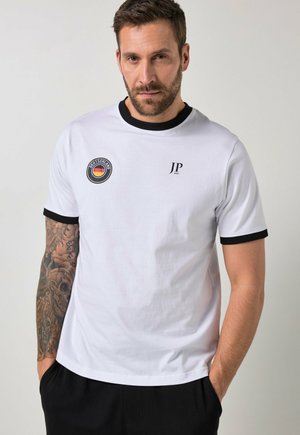 Mann mit tätowiertem Arm, der ein weißes T-Shirt mit schwarzem Rand und Deutschland-Emblem sowie JP 1880-Logo trägt, steht mit den Händen in den Taschen.