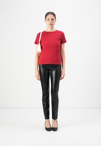 Vrouw staat, draagt een rode T-shirt met korte mouwen, zwarte leren broek, zwarte hoge hakken en draagt een witte schoudertas.