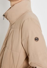 Beige vadderad jacka med hög krage, blixtlås framtill och logopatch på vänster ärm. Slät tyg och strukturerad design.