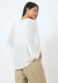 Haut blanc ample à manches longues en tissu texturé à maille ouverte. Présente un col dégagé et des manches drapées. Porté sur un pantalon beige.