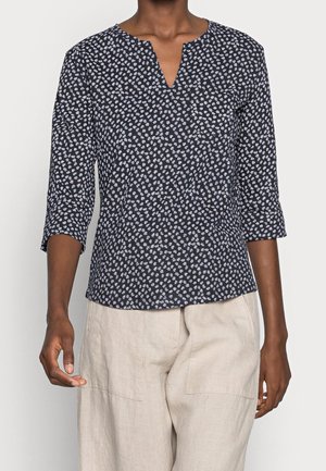Personne portant un haut bleu marine avec de petits motifs floraux blancs et un pantalon beige ample, debout devant un fond clair uni.