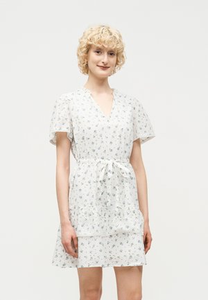 Jeune femme aux cheveux blonds courts et bouclés, portant une robe blanche à motifs floraux avec des manches courtes et une ceinture à la taille, posant devant un fond uni.